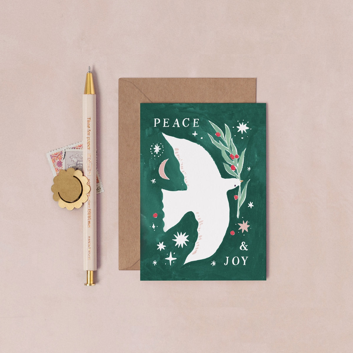 Greeting card with a white silhouette of a bird, stars, and flowers on a green background, with 'Peace & Joy' text, next to a pen and envelope on a beige background.