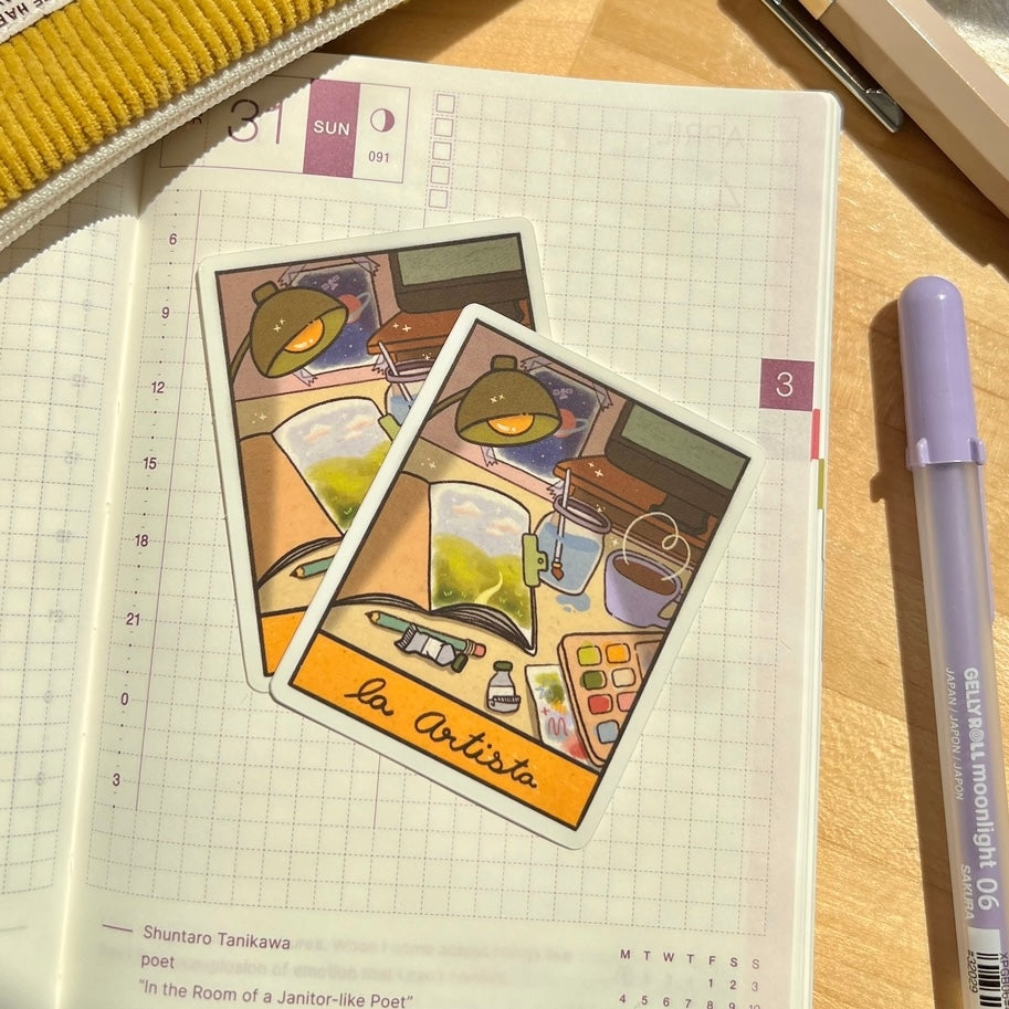 Two illustrated stickers with the text "La Artista" on a notebook with a pen next to them