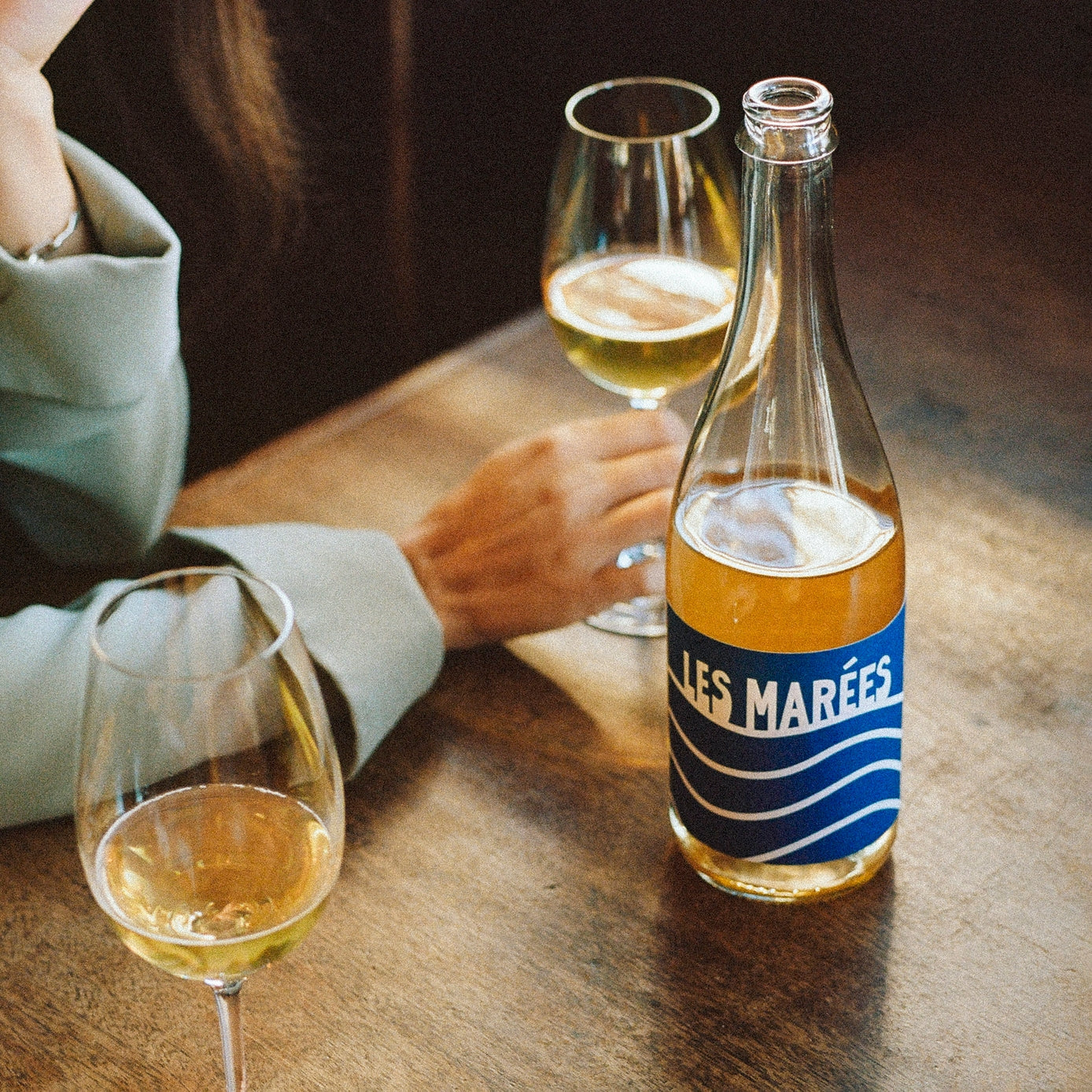Bottle and glasses of 'Les Marées' on a wooden table with a person in the background.