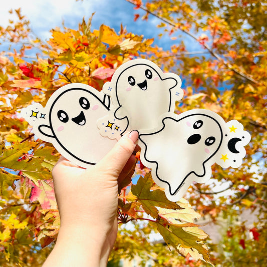 Hand holding three cartoon ghost stickers against a background of autumn leaves