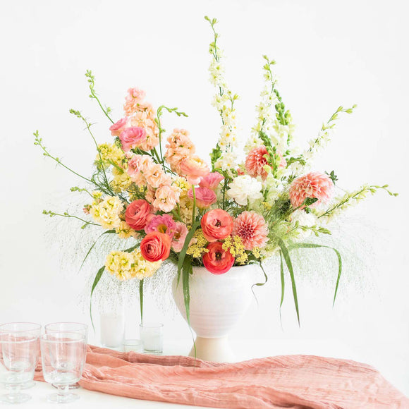 Tall Urn Flower Arrangement in peach, pink, pastel yellow, and white on a table with peach fabric and glasses