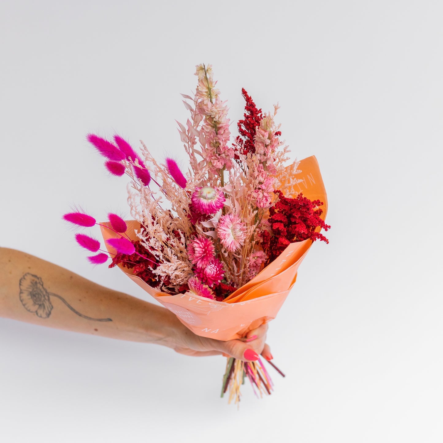 Bouquet of red, pink, and peach dried flowers wrapped in paper held by a person with a tattoo on a light gray background
