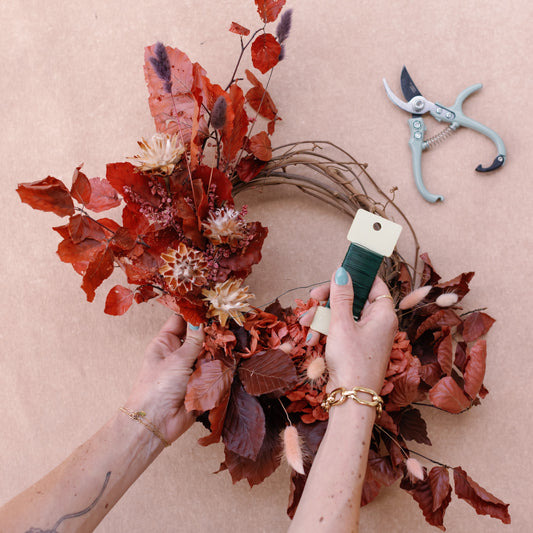 Person crafting a wreath with dried red leaves and flowers on a beige background.