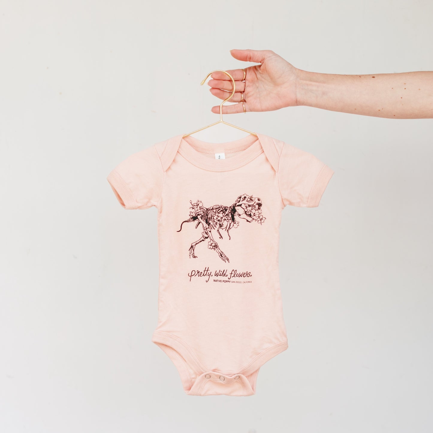 Pink baby onesie with a dino design and text that reads "pretty. wild. flowers." on a hanger against a light gray background