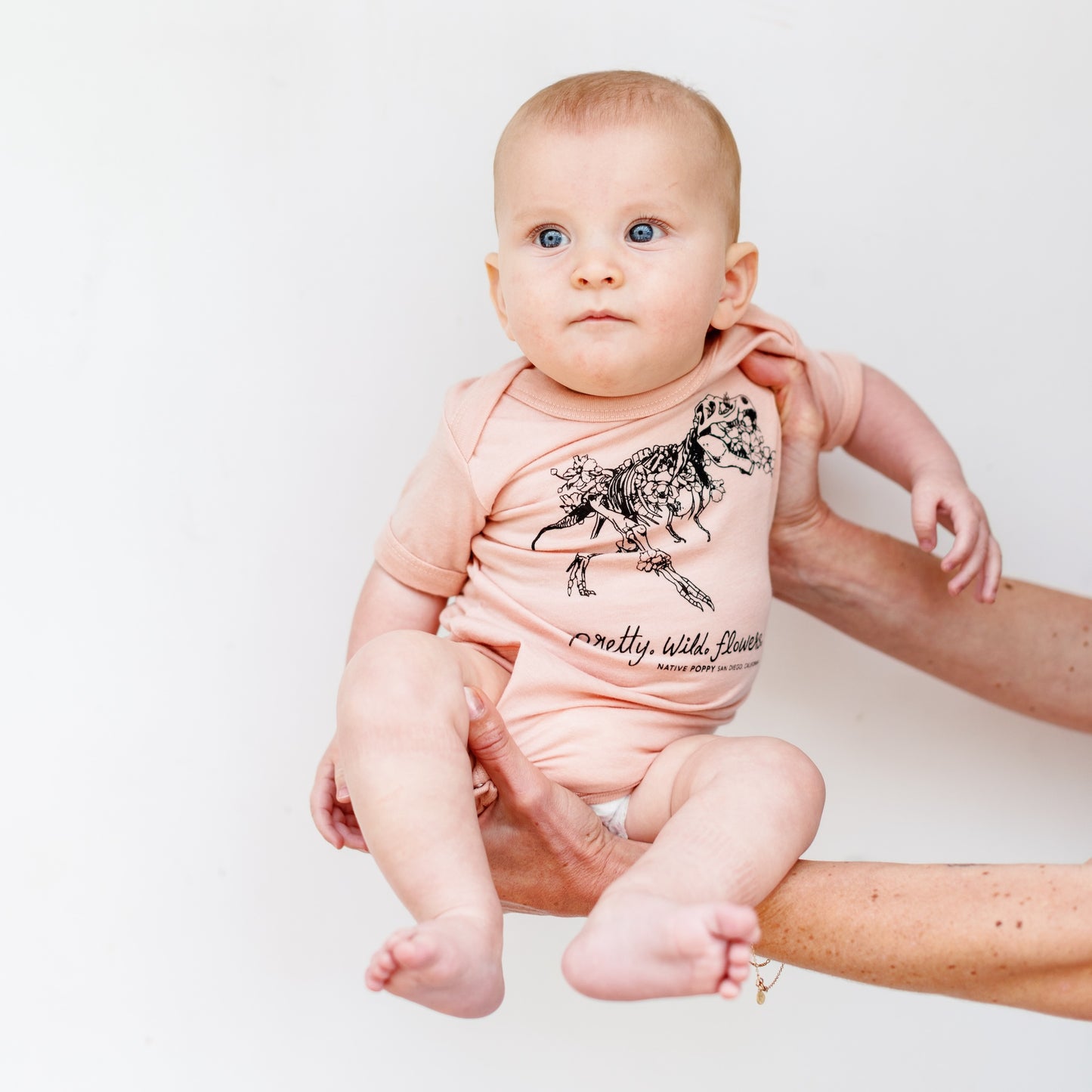 Baby wearing a pink onesie with a dinosaur design and text, held in someone's arms against a white background.