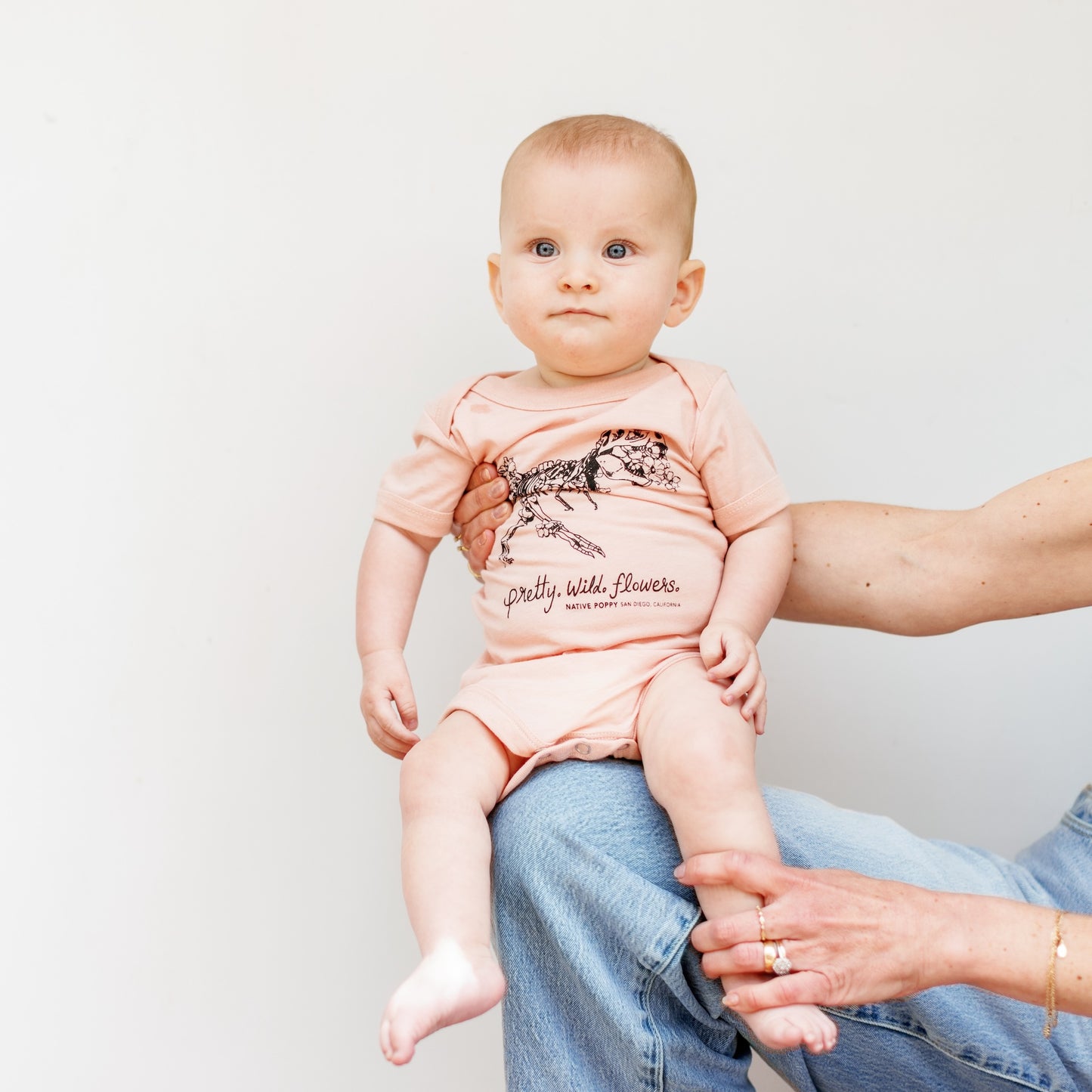 Baby wearing a pink onesie with text and graphics, sitting on a person's lap against a plain background.