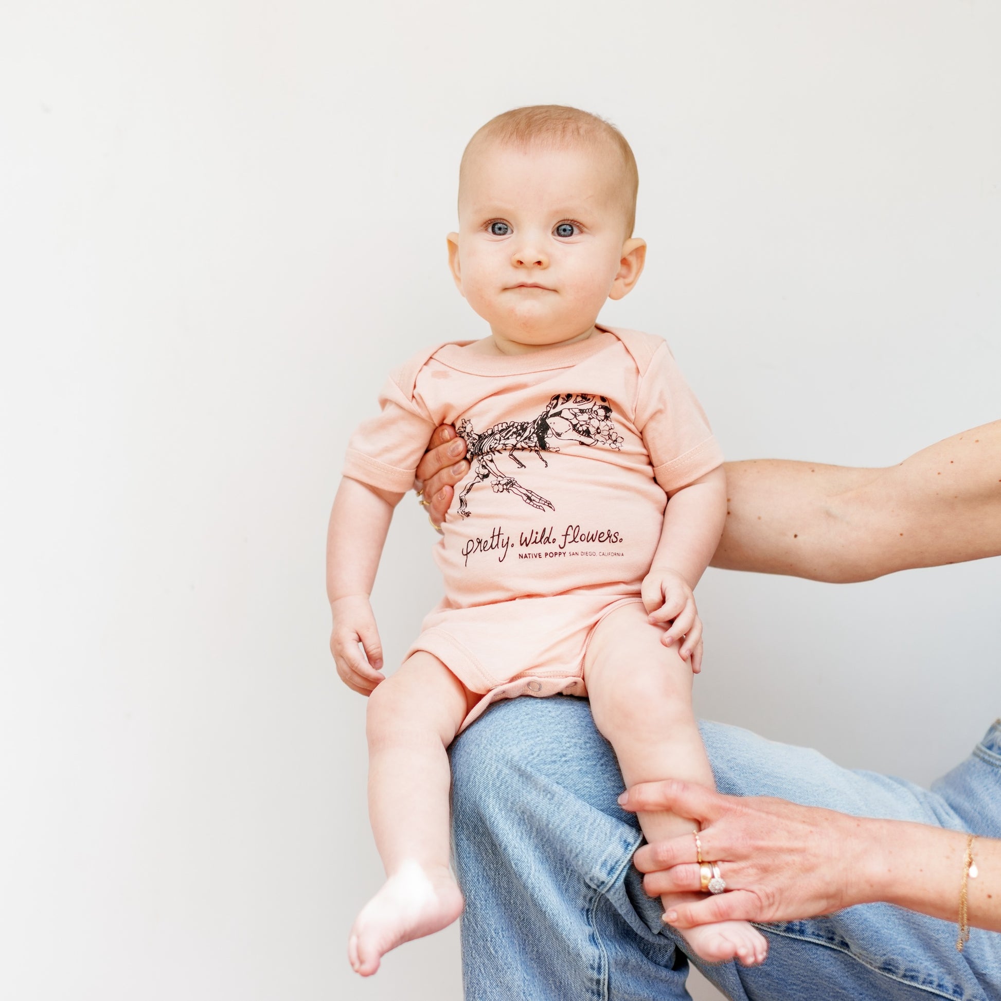 Baby wearing a pink onesie with text and graphics, sitting on a person's lap against a plain background.