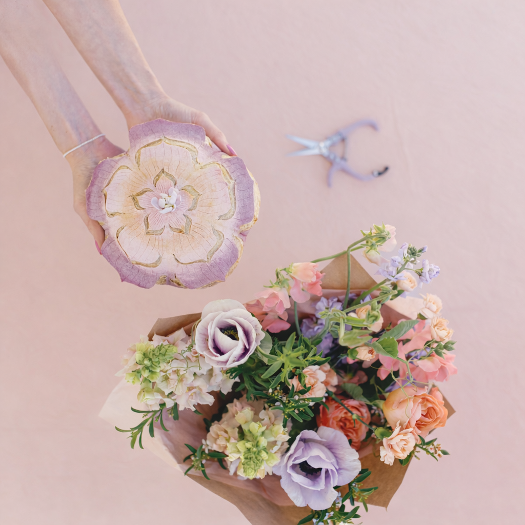 Hand holding a decorative bread with a floral design next to a bouquet of flowers on a pink background