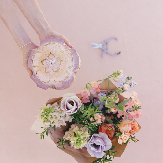 Hand holding a decorative bread with a floral design next to a bouquet of flowers on a pink background