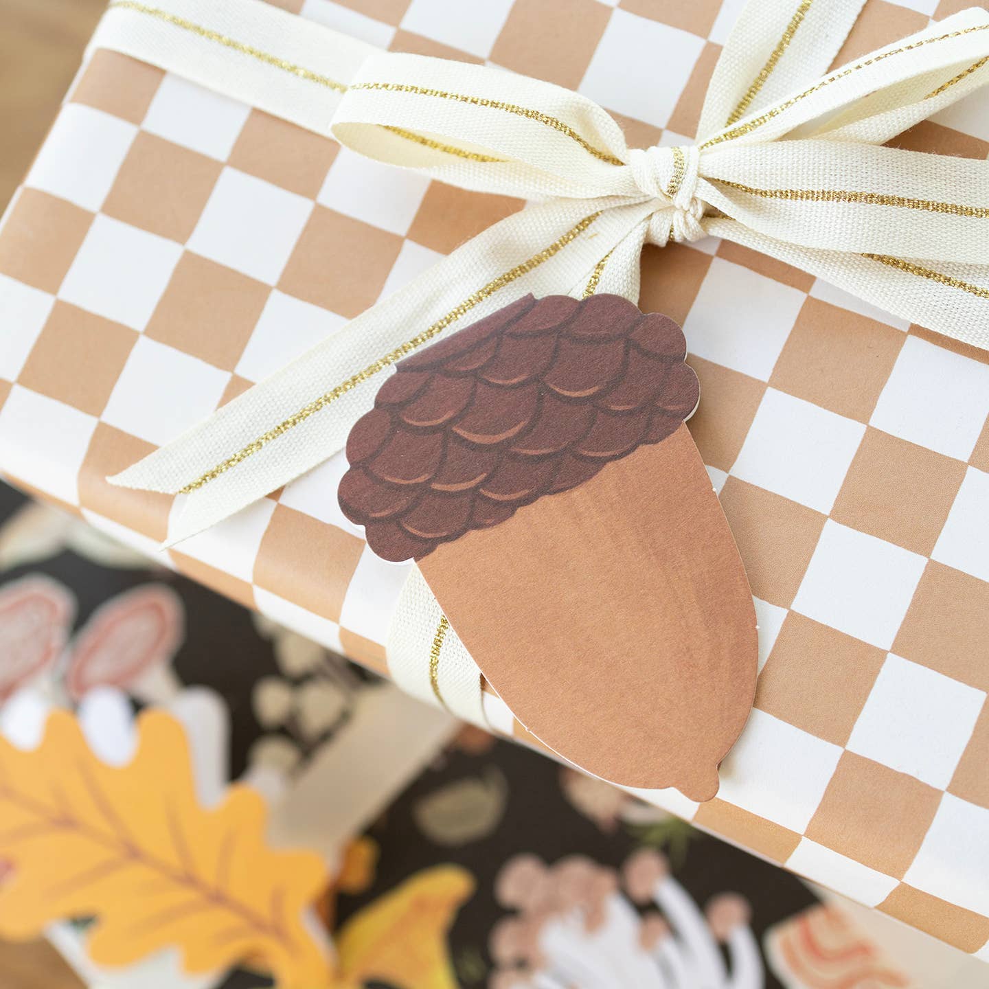 Gift box with checkered paper, white ribbon, and acorn-shaped tag on a blurred autumn-themed background.