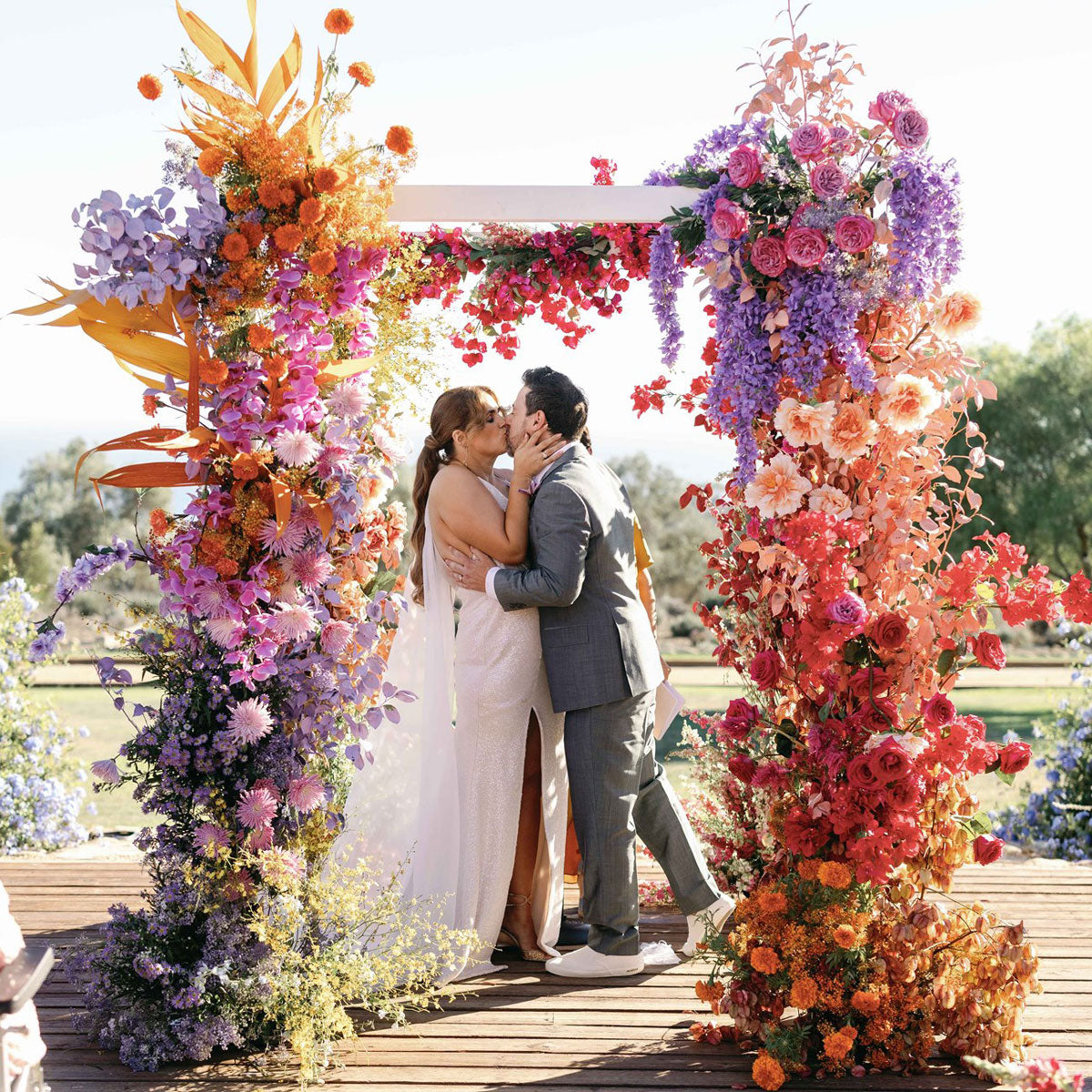 Bride and groom kiss under bright rainbow colored floral wedding chuppah