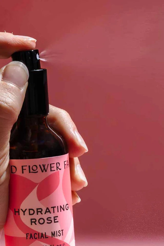 Hand holding a facial mist bottle labeled 'Hydrating Rose' against a pink background