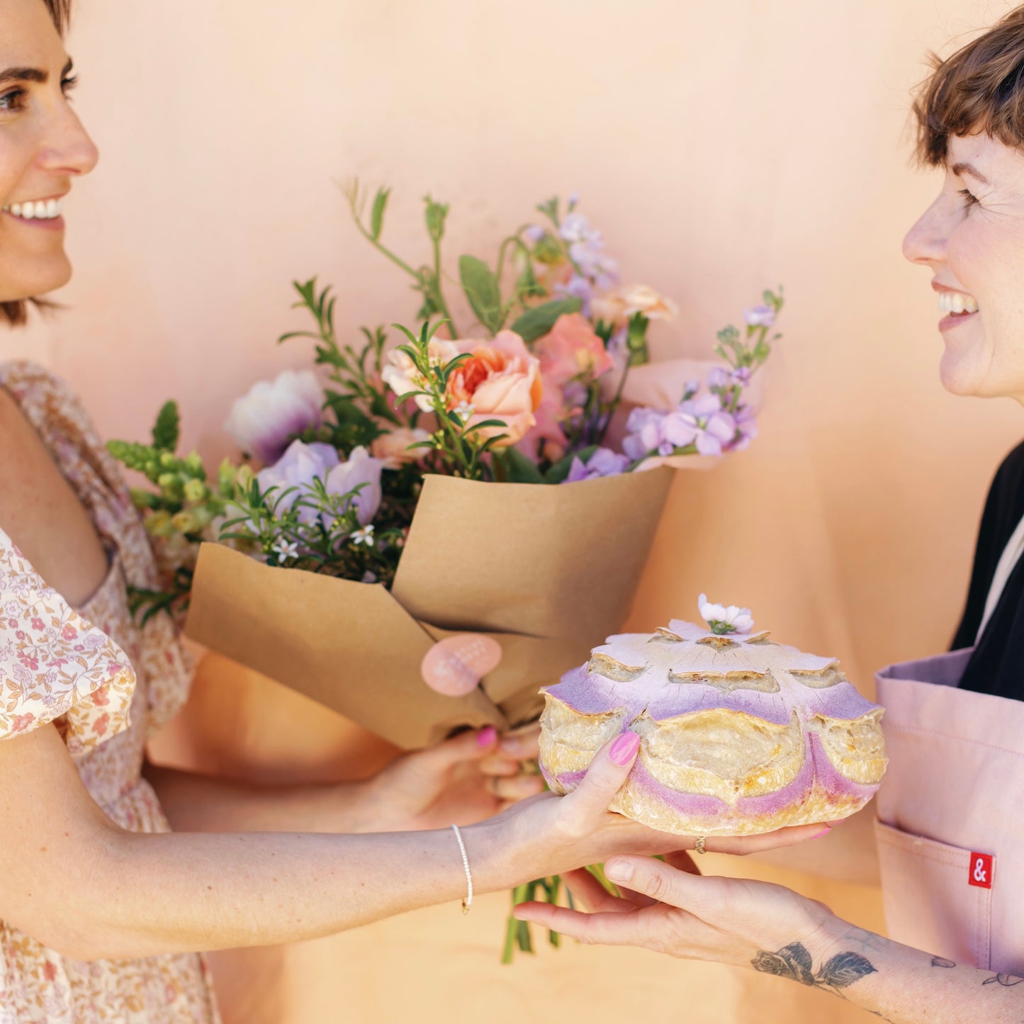 Two women exchanging a decorated loaf and a bouquet of flowers with a soft, warm background.