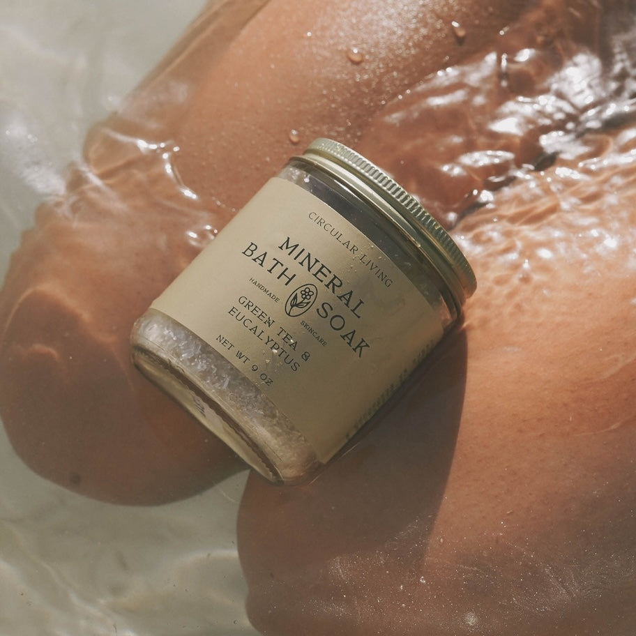 Jar of 'Circular Living' mineral bath soak held over a person's wet skin with water droplets.
