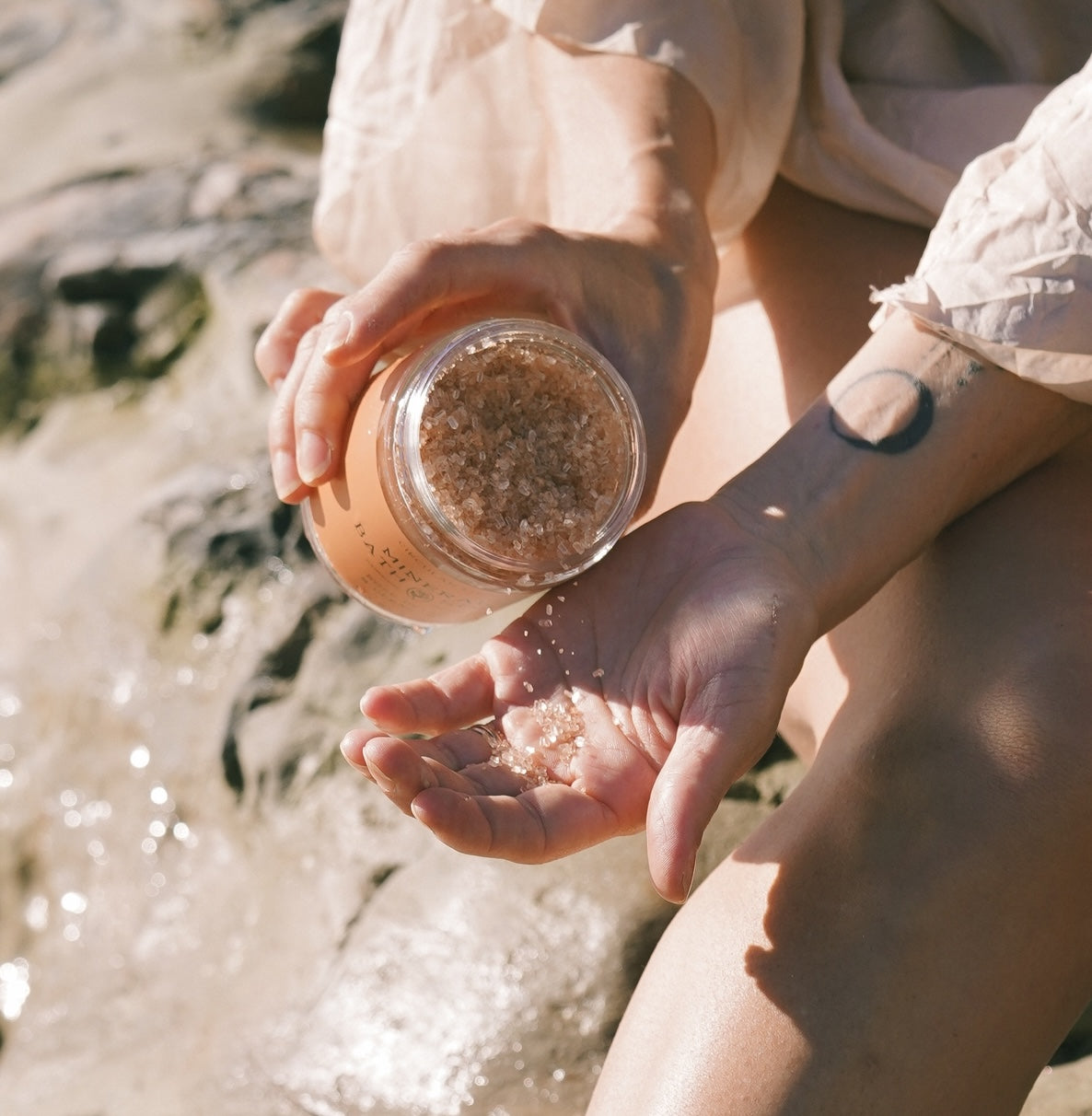 Person holding a jar of mineral bath soak and pouring it into their hand by a body of water.