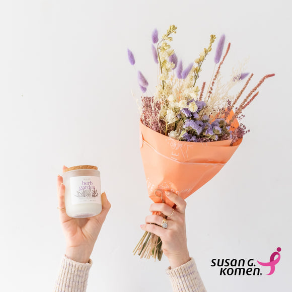 Arms outstretched holding dried flower bouquet and Mama's Herb Garden candle and the Susan G Komen pink ribbon logo