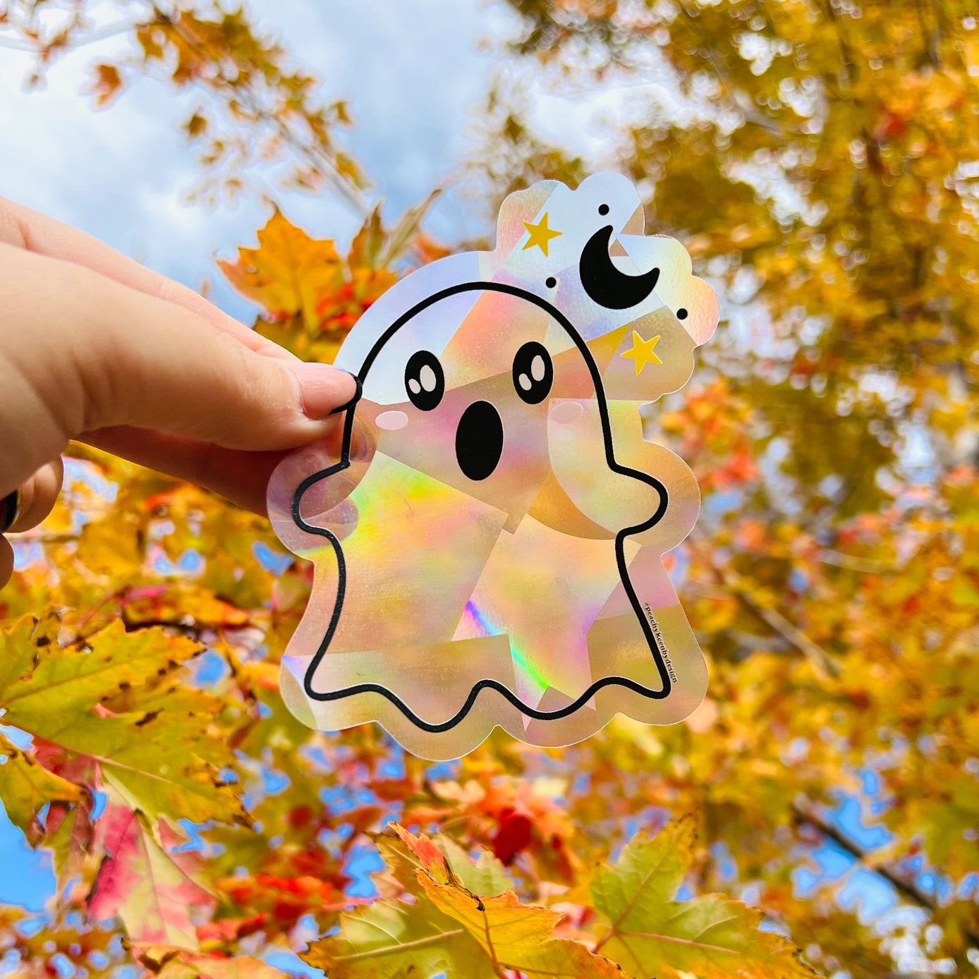 Holographic ghost sticker held by a hand with autumn leaves in the background