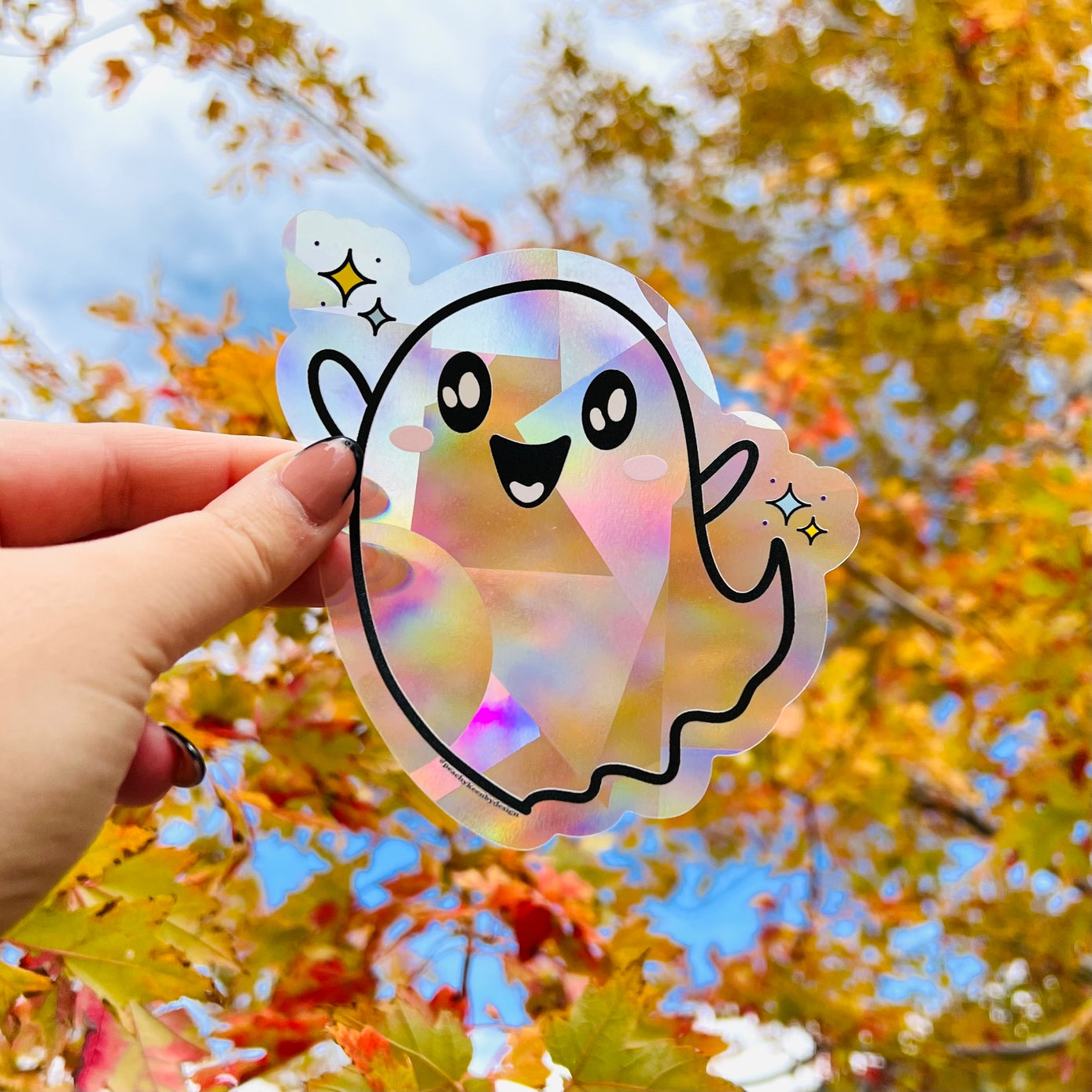 Hand holding a colorful ghost-shaped sticker with autumn leaves in the background