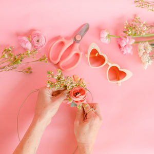 Crafting a flower crown with pink and white flowers, next to pink snips and heart glasses