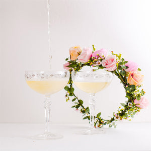 Pink and peach rose floral crown displayed on two coupe glasses being filled with champagne